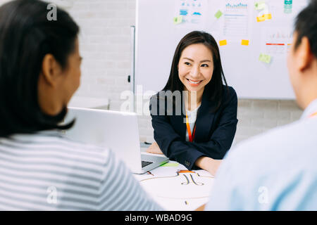 Asian mature business woman smiling and looking at other business people while sitting in meeting. Smart middle age lady having discussion. Stock Photo