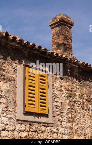 Detail of a house made of stones. Frame of the window and a yellow closed shutter. Roof with chimney made of bricks. Blue sky, light clouds. Sibenik, Stock Photo