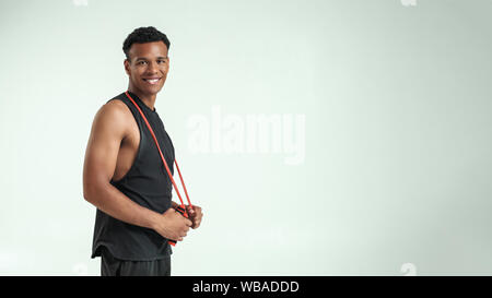 Feeling happy. Cheerful and young afro american man in sportswear holding a jumping rope at his shoulders and looking at camera with smile while standing against grey background. Sport equipment. Active lifestyle. Web banner Stock Photo