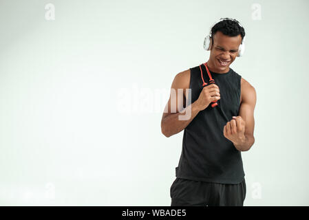 I can do everything. Cheerful young afro american man in headphones holding a jumping rope, smiling and clenching a fist while standing against grey background. Sport equipment. Motivation Stock Photo