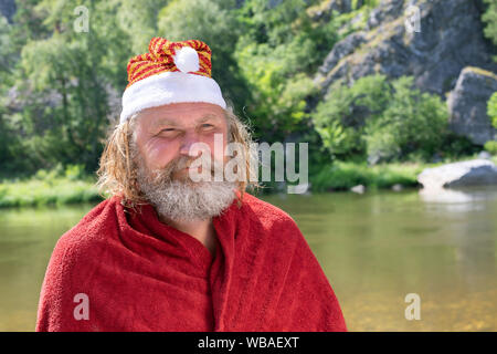 charismatic elderly man with a beard in a Santa Claus hat and red coat smiling. Against the background of a summer landscape, river, mountains, forest Stock Photo