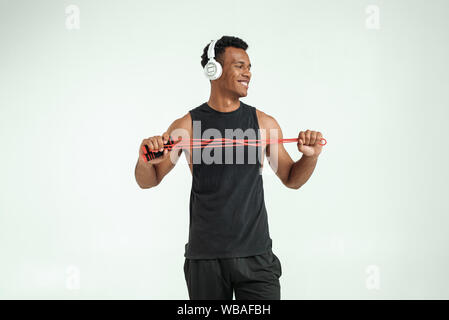 Exercising with music. Cheerful and sporty young afro american man in headphones holding a jumping rope and smiling while standing against grey background. Sport equipment. Active lifestyle Stock Photo