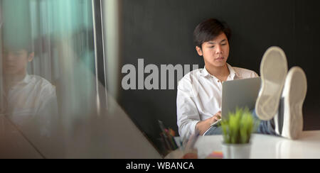 Young male freelancer put his feet on the table while working on a laptop computer in trendy work space Stock Photo