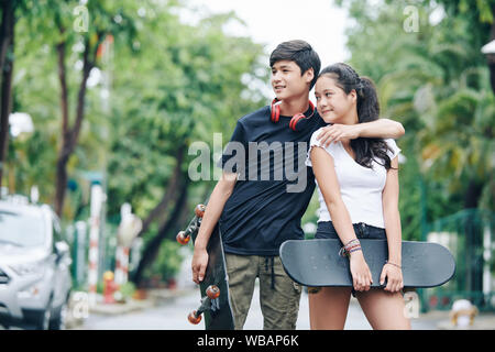 Cheerful hugging mixed-race brother and sister standing outdoors with skateboards in hands Stock Photo
