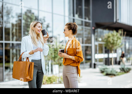 Two young businesswomen talking near the office building, having a small talk during the coffee break outdoors. Stock Photo