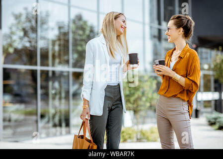 Two young businesswomen talking near the office building, having a small talk during the coffee break outdoors. Stock Photo