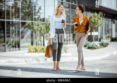 Full body portrait of a two young businesswomen walking with coffee cups near the modern office building outdoors Stock Photo
