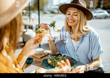 Two female best friends eating healthy food while sitting together on a restaurant terrace on a summer day Stock Photo