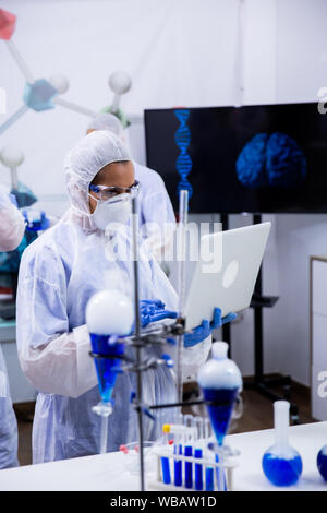 Scientist typing on her computer data from exeperiments with smoking blue solution in test tubes. Stock Photo