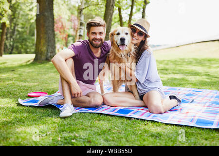 Cheerful young couple sitting on a blanket with their dog while out in the park together enjoying the nature Stock Photo