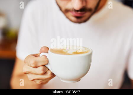 Close up of coffee with beautiful latte art being held by young man in cafeteria Stock Photo