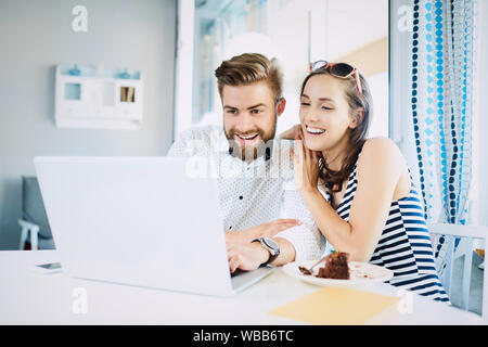 Cheerful young couple looking at laptop laughing and talking while sitting in stylish bright restaurant Stock Photo