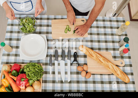 Hands of woman mixing salad when her boyfriend cutting cucumber on dinner table, view from above Stock Photo