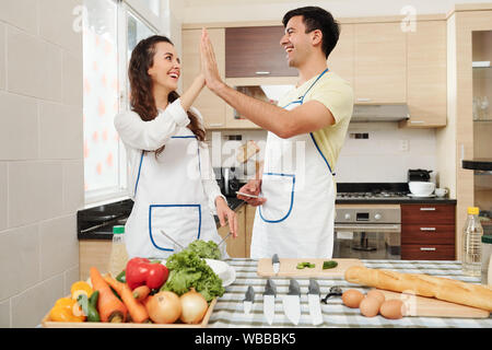 Happy young couple standing at kitchen table in aprons and giving each other high five Stock Photo