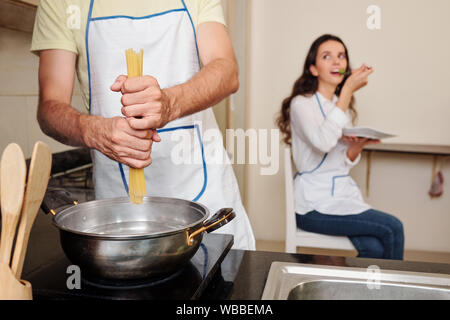 Young man putting spaghetti in boiling water when his wife eating salad in background Stock Photo