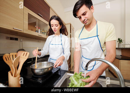 Young man rinsing salad ingredients under tap water while his wife cooking pasta for dinner Stock Photo