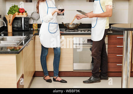 Cropped image of young woman drinking wine while her husband reading recipe on tablet computer and stirring vegetables in frying pan Stock Photo