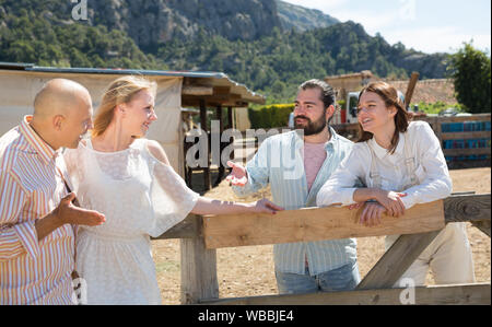 Happy adult friends breezily chatting near wooden fencing in country house in warm spring day Stock Photo