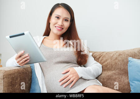 Portrait of lovely young Vietnamese woman sitting on sofa with digital tablet touching her baby bump and smiling at camera Stock Photo