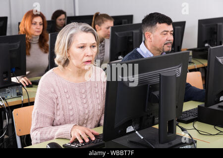 Computer lessons for adults. Mature woman learning to use computer in classroom with group Stock Photo