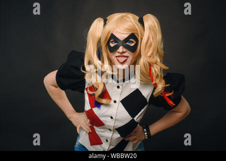 A girl in a suit and wig on a black background is smiling and showing tongue. Girl cosplay character and having fun in the studio on Halloween. Stock Photo