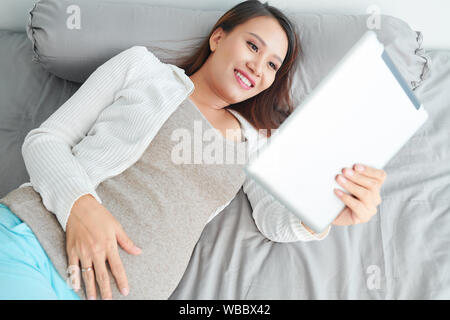 Happy young beautiful Vietnamese woman lying in bed and watching funny videos on digital tablet Stock Photo
