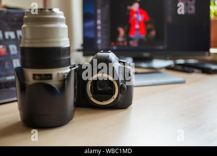 Professional photo studio equipment on desk.Big black dslr photocamera and long white zoom lens.Computer screen with retouched images on background.Co Stock Photo