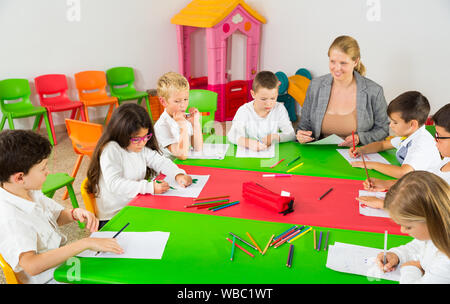 Friendly female teacher talking to children, sitting together around desk in classroom Stock Photo