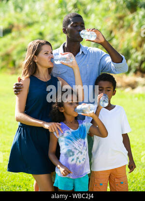 Happy mixed race family with two children enjoying walk on green meadow, drinking clear water Stock Photo