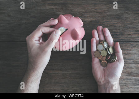top view of man holding coins in one hand and putting money in piggy bank with other hand, saving money concept Stock Photo