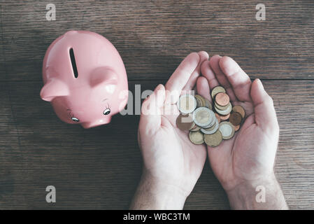 top view of hands of man holding coins next to piggy bank on rustic wooden table, saving money concept Stock Photo