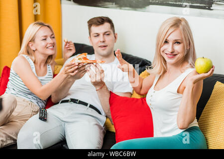 Beautiful caucasian sporty woman eating apple while her friends enjoying pizza, doing stop sign, smiling looking at camera. Dieting concept. Healthy F Stock Photo