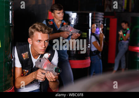 Young positive cheerful glad guy holding colored laser guns and took aim during laser tag game in labyrinth Stock Photo
