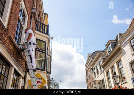 Utrecht, Netherlands - JULY 01, 2019: From below side view of male worker in white uniform standing on ladder cleaning window of old building Stock Photo