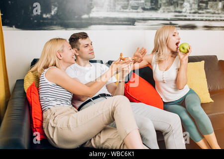 Fast food or healthy food. Caucasian blonde lady choosing between apple and pizza slice, doing stop sign. Sporty woman eating fruit while her friends Stock Photo
