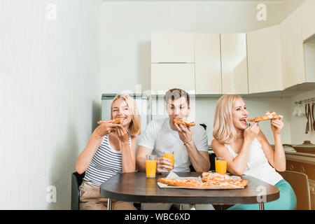 Friendship, people and food concept. Three caucasian happy students eating pizza in kitchen.They are having party at home, laughing and having fun, dr Stock Photo
