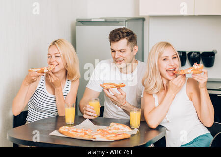 Two happy beautiful blonde caucasian young women with their friend enjoying tasty pizza and drinking fruit smoothies, sitting at table in kitchen, tal Stock Photo