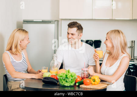 Students relaxing in kitchen. Three friends enjoying fresh juice, having fun together in morning, laughing loudly. Lifestyle, friendship and healthy f Stock Photo