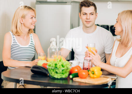 Friends relaxing in kitchen.Three caucasian students sitting at table, thinking what to cook for dinner, drinking smoothies and talking. People, healt Stock Photo