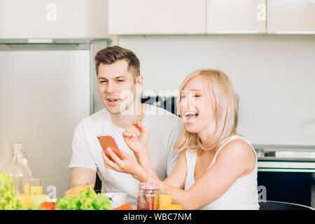 Beautiful young caucasian couple sitting in kitchen, looking at mobile phone together, watching funny video and laughing. Joyful blonde woman and hand Stock Photo