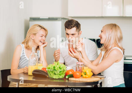 Cheerful best friends viewing funny photos, watching video in social networks via smartphone and laughing while cooking in kitchen. Three young friend Stock Photo