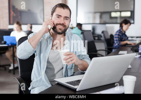Smiling young Hispanic man talking on phone in busy modern open plan office Stock Photo