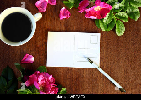 top view of blank white postcard, a pen, coffee and wild roses on wooden table background. romantic composition of pink and brown warm and earthy colo Stock Photo