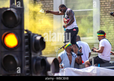 Powder is thrown by a man riding on top of a sound system truck. The main events of Notting Hill Carnival 2019 got underway on Sunday, with over a million revellers hitting the streets of West London, amongst floats, masqueraders, steel bands, and sound systems. Stock Photo