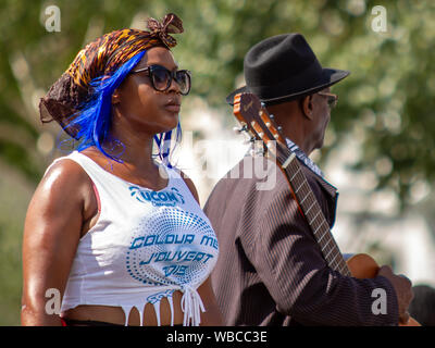 A woman from the United Colours of Mas (UCOM) band, beside an older man with a Spanish guitar. The main events of Notting Hill Carnival 2019 got underway on Sunday, with over a million revellers hitting the streets of West London, amongst floats, masqueraders, steel bands, and sound systems. Stock Photo