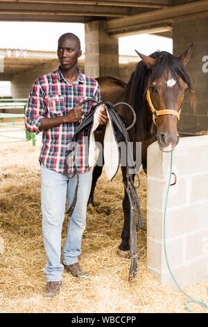 Male african american farmer removes saddle from horse at stable Stock ...