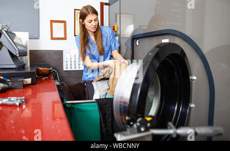 Efficient woman in uniform taking out clothes from washing machine at laundry Stock Photo