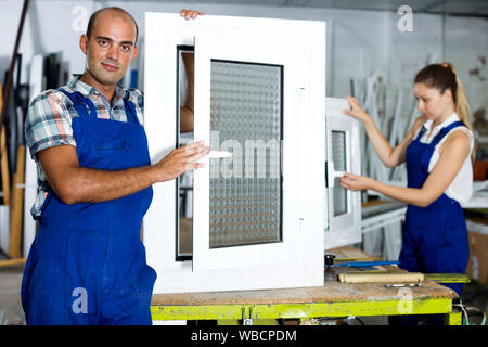 Positive workman in blue overalls demonstrating pvc window in workshop Stock Photo