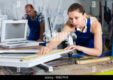Young workwoman is checking dimensions of finished plastic window using measuring tape at factory. Stock Photo