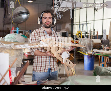 Cheerful male aircraft enthusiast in aviator headphones holding sports airplane models in workshop Stock Photo